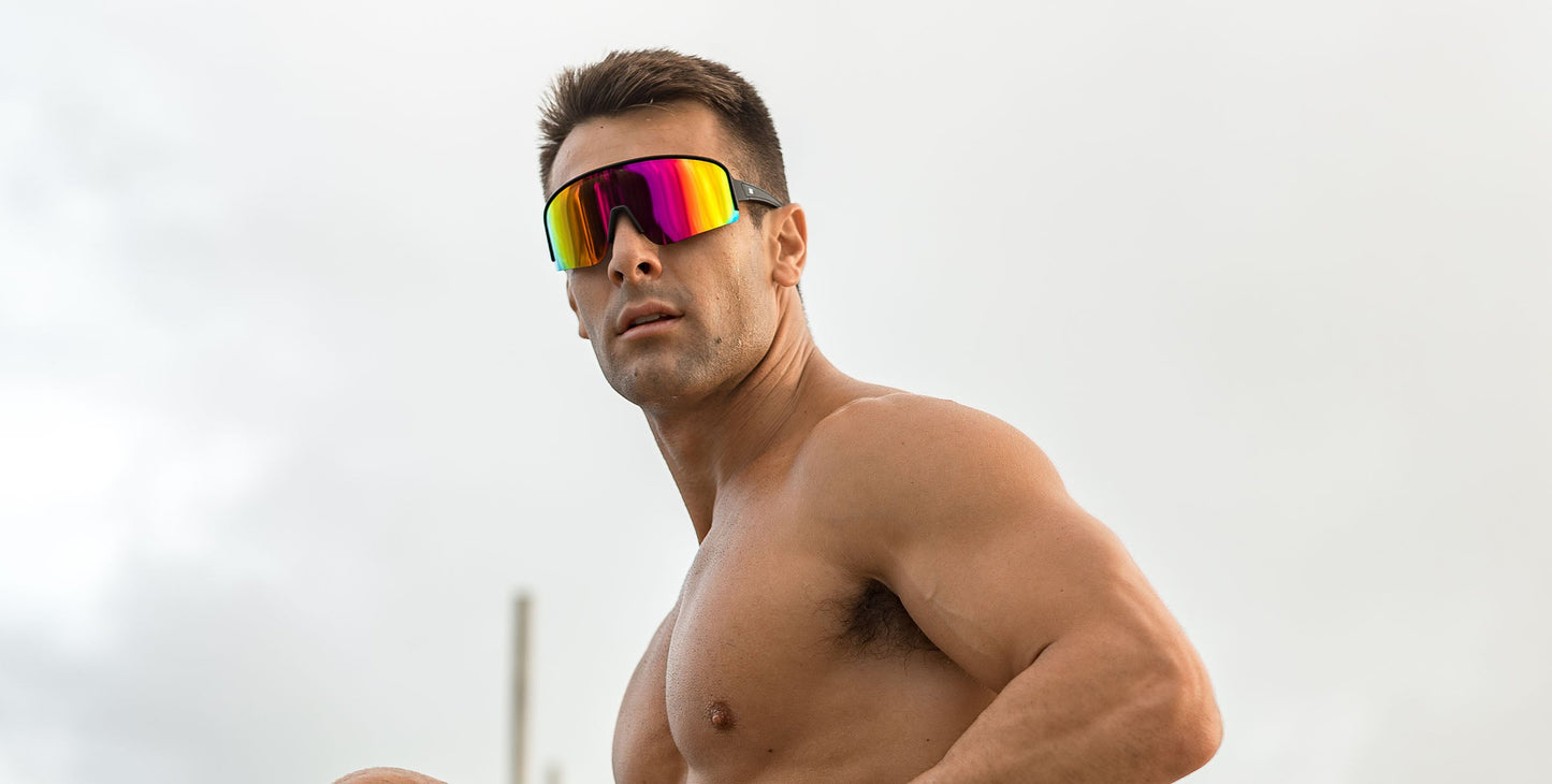 A shirtless, muscular man with short hair stands outdoors under a cloudy sky, wearing large, reflective FLL SND sunglasses from Neven Eyewear and gazing to the side with a serious look, his pose accentuating his defined physique.