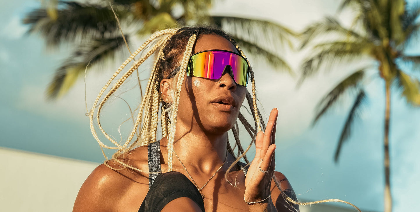 A woman with long blonde braids, dressed in athletic attire and wearing Neven Eyewear FLL SND sunglasses, exercises outdoors, raising her knee and hand under a warm sun with blurred palm trees and blue sky behind her.