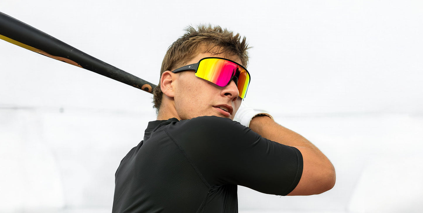 A young man in a black shirt and Neven Eyewear’s FLL SND sunglasses holds a baseball bat, glancing over his shoulder with one hand gloved, set against a vibrant, blurred background.