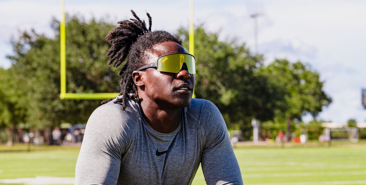 A man with dark skin and dreadlocks wears a gray athletic shirt and Neven Eyewear PR polarized sunglasses. On a football field with green trees, he looks focused behind his large, reflective shades.