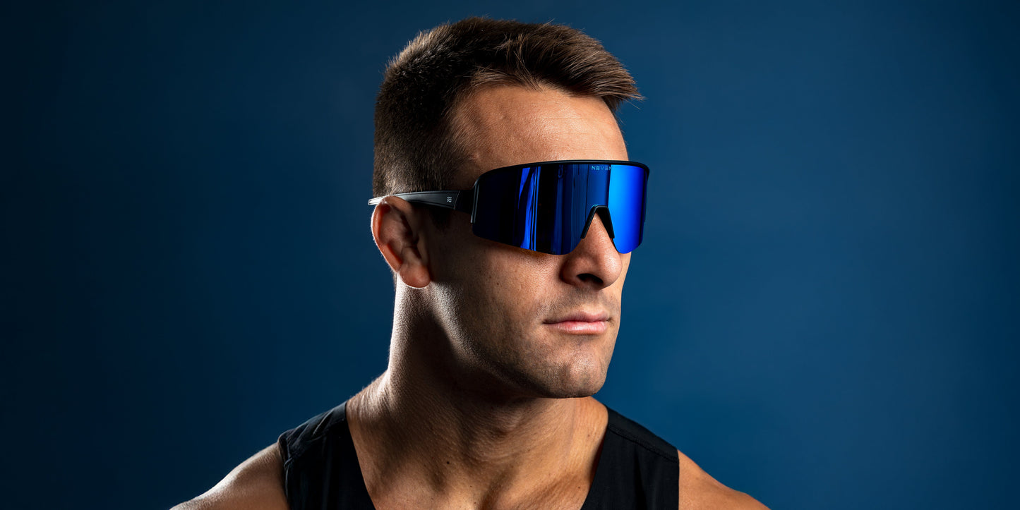 A man with short brown hair in a black sleeveless shirt wears Neven Eyewear Strides sunglasses and looks confidently to the right against a solid dark blue background.
