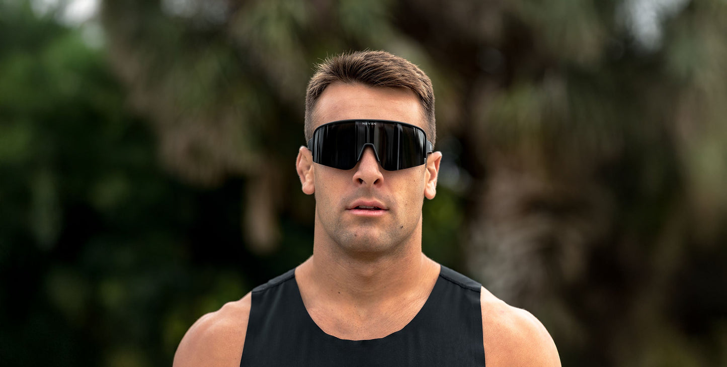 A man with short brown hair faces forward, wearing large Neven Eyewear Threshold sunglasses with polarized UV400 protection and a sleeveless black top, set against a blurred backdrop of green foliage and tree trunks.