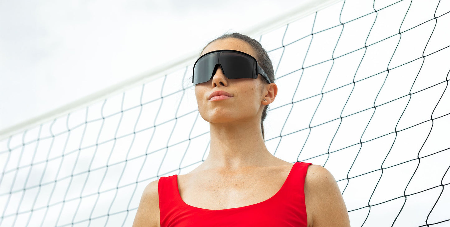 A woman in a red tank top stands confidently by a volleyball net, wearing Neven Eyewear's Threshold sunglasses with polarized UV400 protection. The bright, cloudy sky enhances the summery, sporty vibe of the scene.