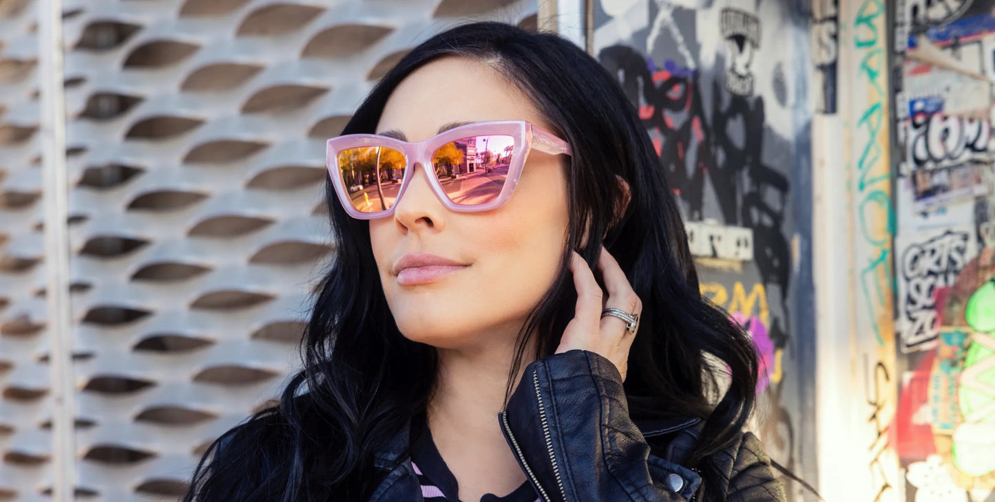 A confident woman with long black hair wears Neven Eyewear's Eclipse shiny pink cat-eye sunglasses and a black leather jacket near a graffiti wall. An urban scene reflects in her glasses as she touches her neck and smiles slightly.