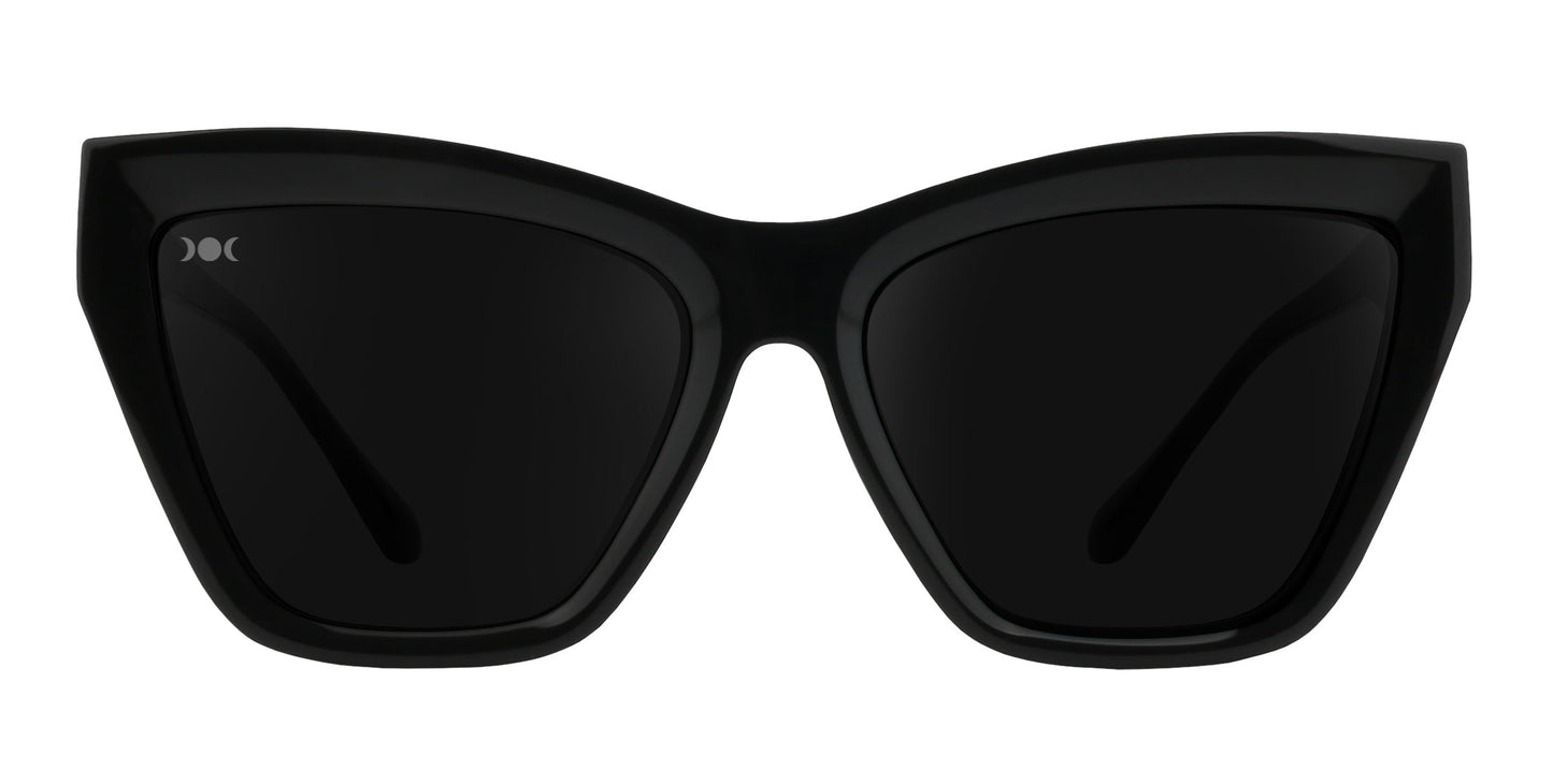 Front view of Nocturnal by Neven Eyewear: black statement sunglasses with thick, glossy cat-eye frames, dark tinted lenses, a slight upward flair at the edges, and a small white Neven logo on the upper left lens.