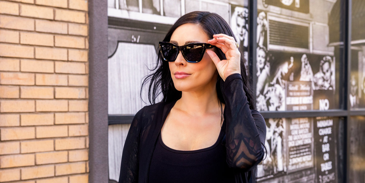 A woman with long dark hair wears Neven Eyewear's Nocturnal black statement sunglasses and a black outfit, posing by a brick wall with bold black-and-white street art, lightly touching her sunglasses.