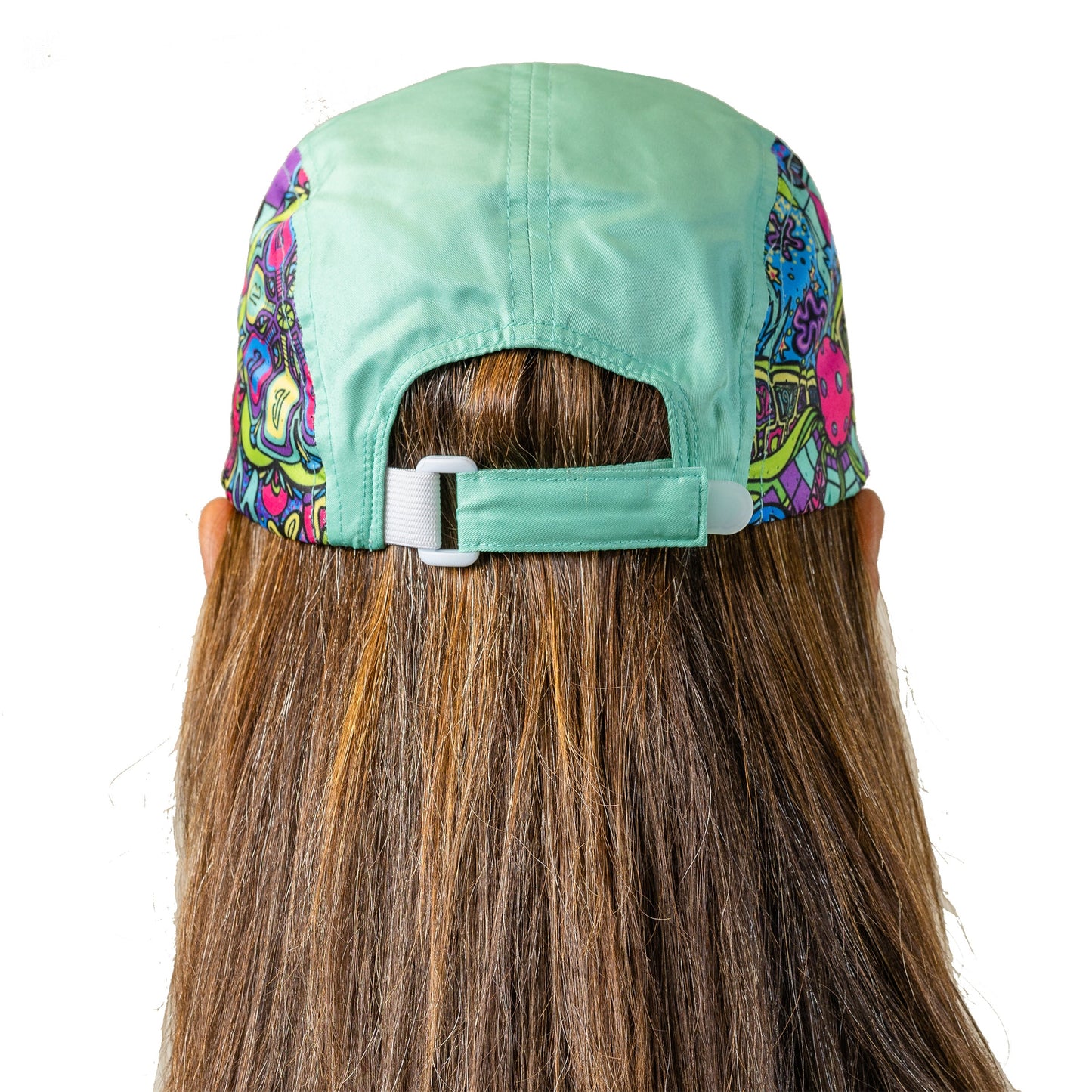 A person with long brown hair is shown from behind, wearing the Pickleball Bella PB Cap Groovy—a green cap with colorful 70’s-inspired side panels and an adjustable strap and buckle. The background is plain white.
