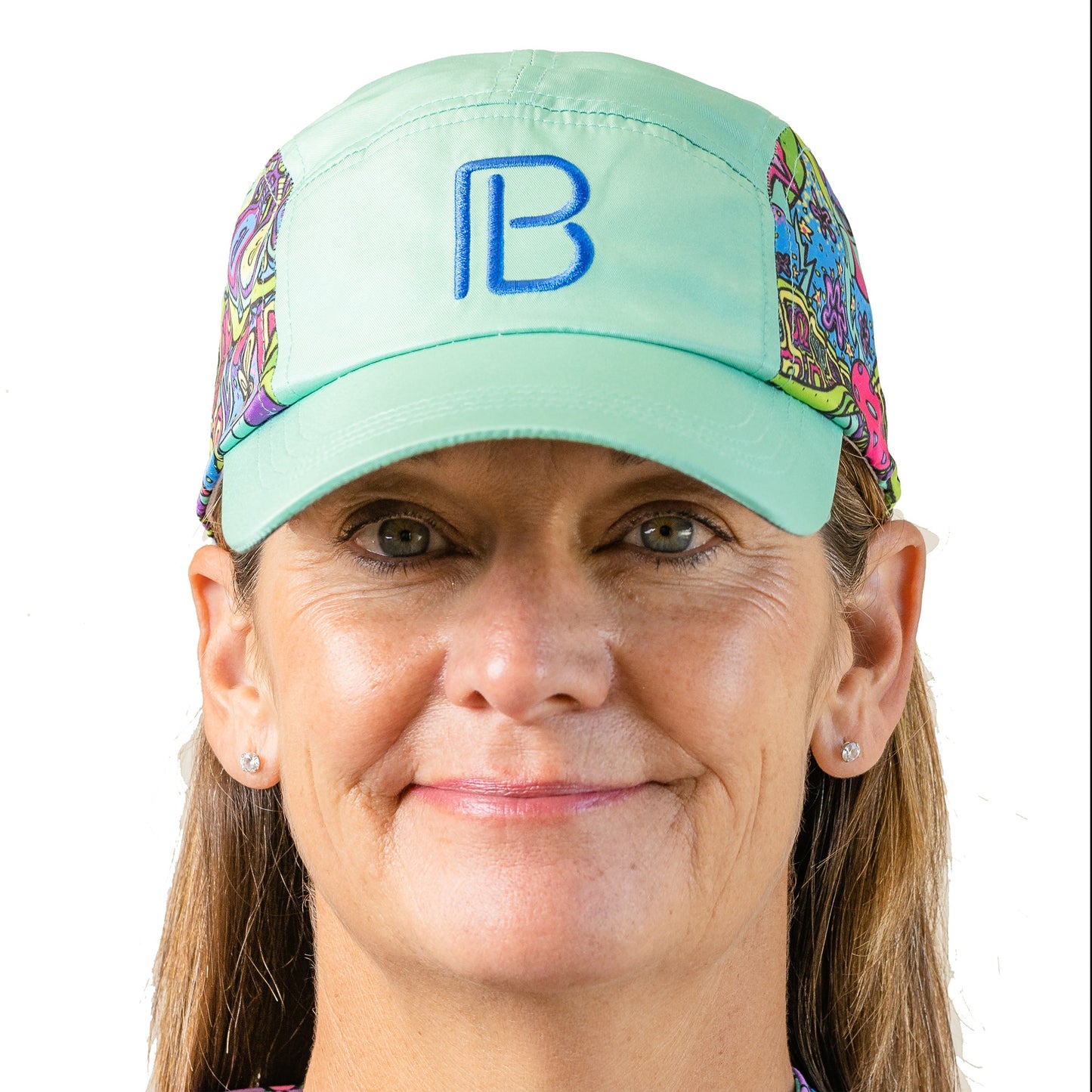 A woman with light brown hair wears the Pickleball Bella PB Cap Groovy, featuring a green base, blue "B" logo, and colorful patterned sides. She smiles slightly at the camera with small stud earrings against a plain white background.