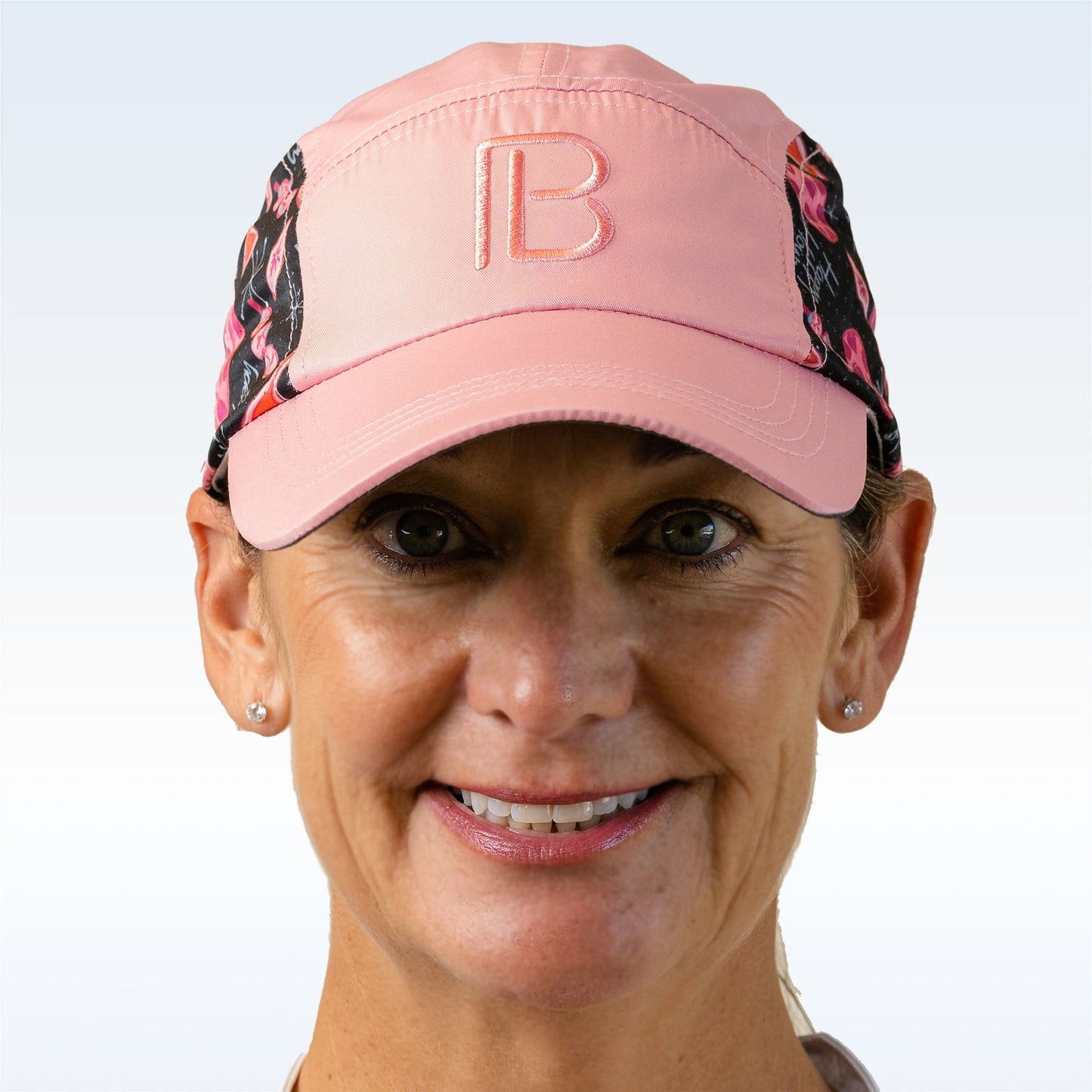 A smiling middle-aged woman wears the Pickleball Bella PB Cap Martini 2-4-1 Happy Hour with a "B" logo, black and pink patterned side panels, and an adjustable velcro strap—perfect for pickleball—plus stud earrings and subtle makeup.