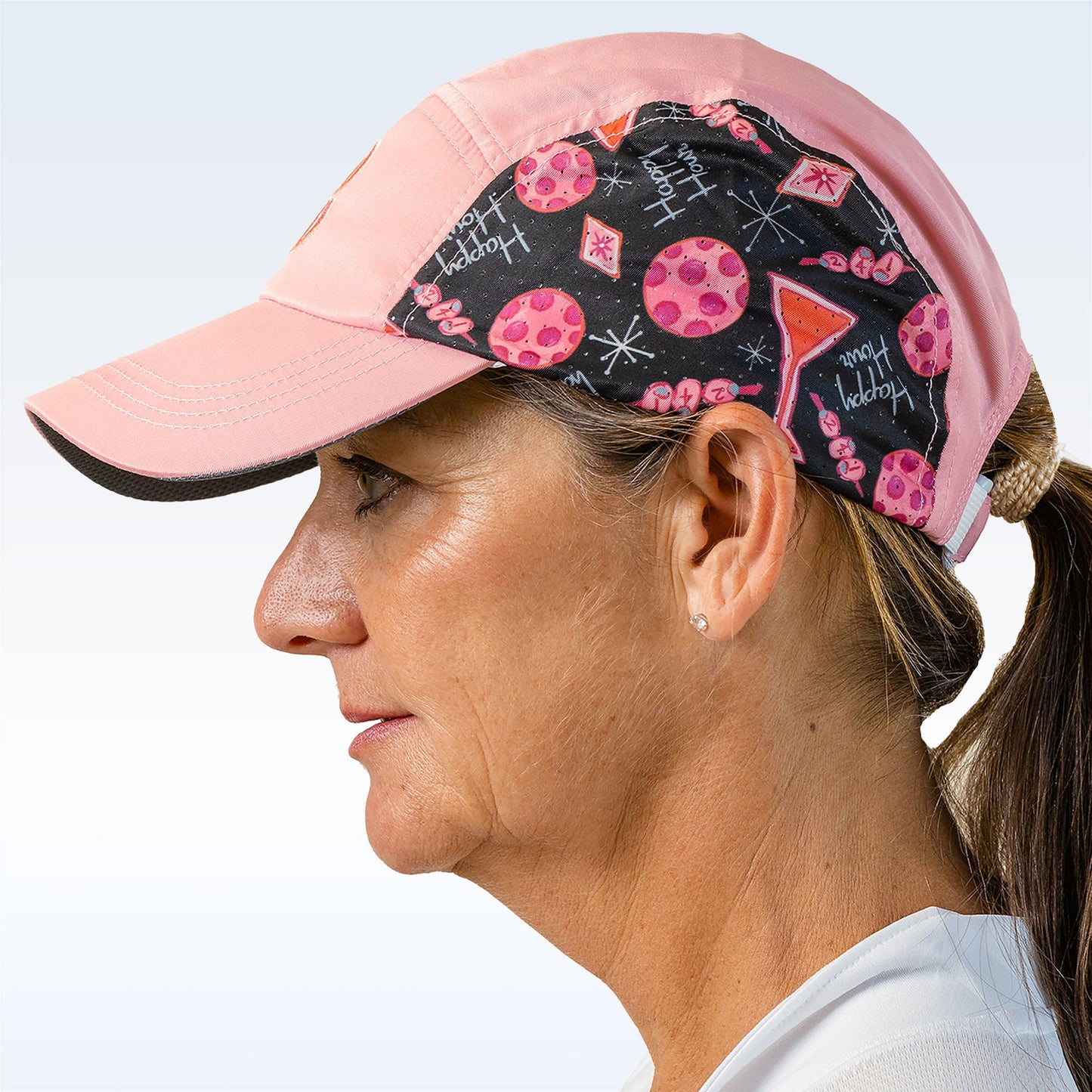 A woman in profile models the Pickleball Bella PB Cap Martini 2-4-1 Happy Hour, featuring a whimsical black panel with words, cocktail glasses, and pink dots. Her brown hair is in a low ponytail, and the adjustable cap secures with velcro.