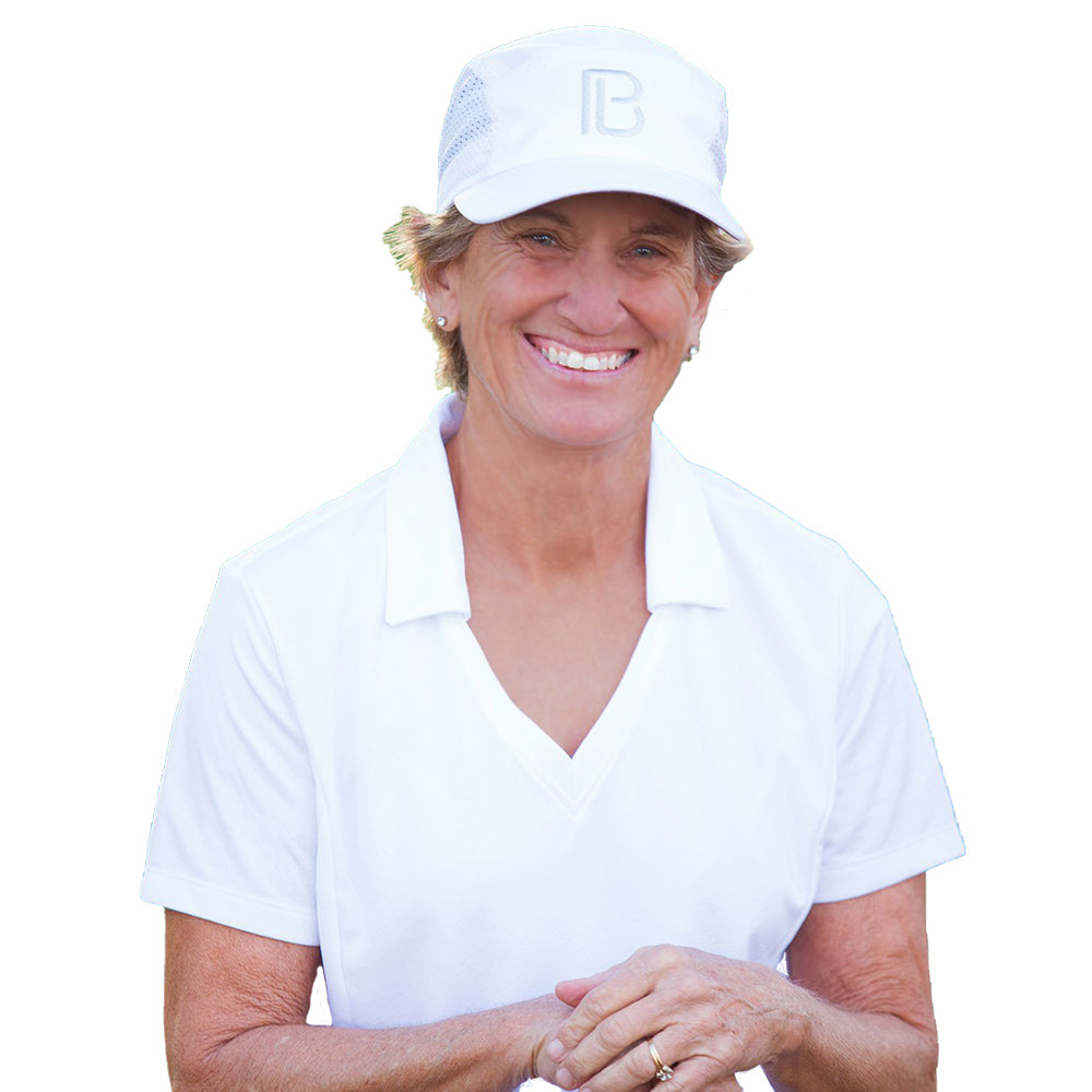 A smiling older woman stands against a white background, wearing a Pickleball Bella PB Cap in white and a collared shirt. She has short hair, small hoop earrings, and her hands are clasped in front of her.