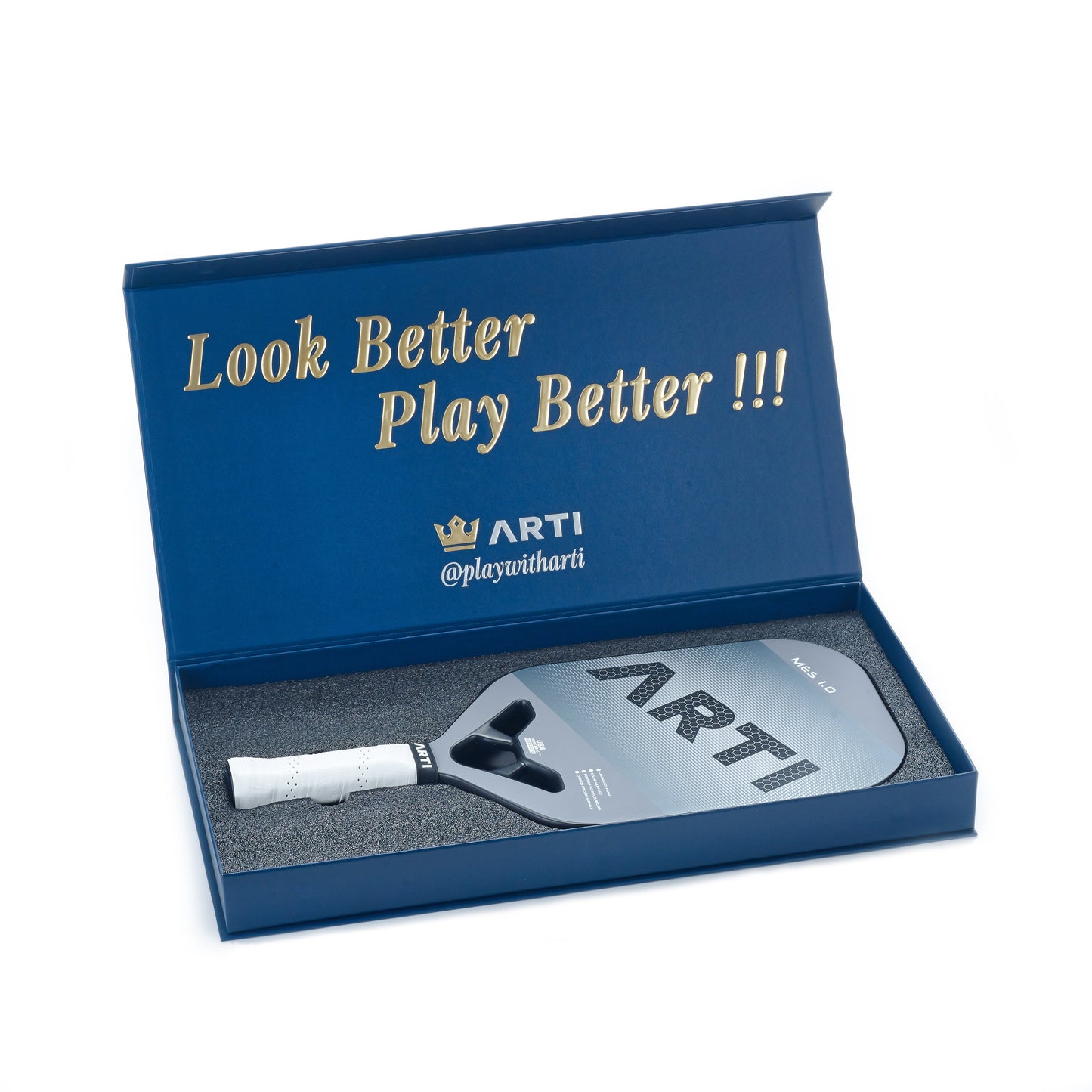 A blue gift box with gold "Look Better Play Better!!!" text holds an ARTI Carbon Fiber "MASTERY ELITE 1.0" USAPA-approved pickleball paddle (gray T700 carbon fiber, white grip, custom-cut foam); lid displays ARTI logo and @playwitharti in white.