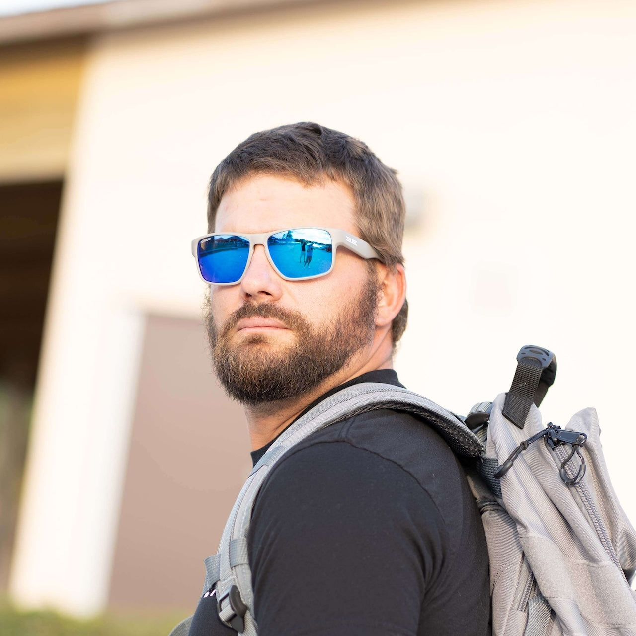 A bearded man with short brown hair and a gray backpack glances back, wearing Epoch Eyewear's Charlie Polarized Sunglasses with reflective blue lenses. He stands in front of sunlit buildings, ready for adventure.