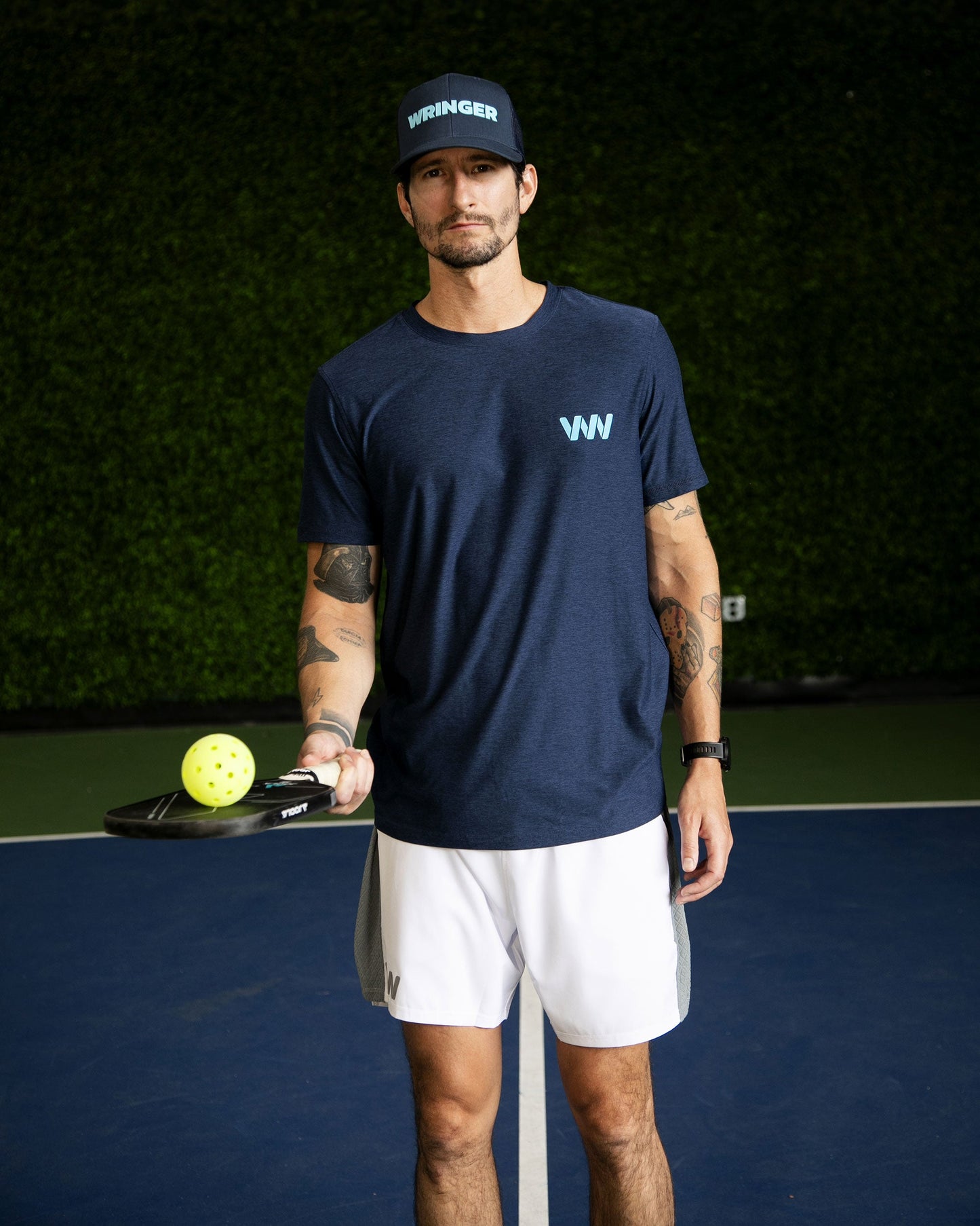 A man stands on a blue pickleball court holding a paddle and yellow ball. He wears a navy WRINGER hat, navy WVN t-shirt, Mojave Performance Shorts 6" Lined by Wringer Wear, and has several tattoos. A green hedge wall is behind him.