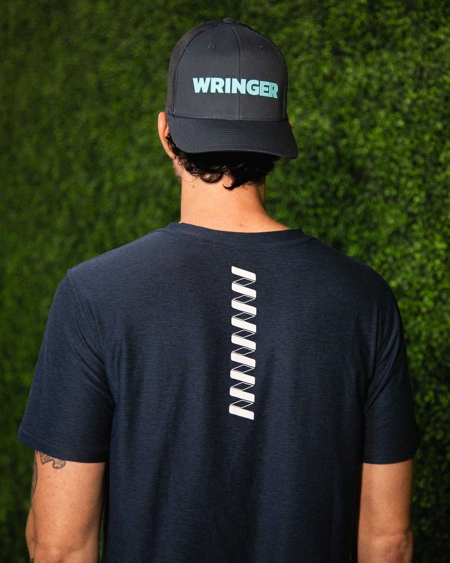 A person stands with their back to the camera, wearing a dark T-shirt with a white zigzag and an Adjustable Mesh WRINGER Hat by Wringer Wear, featuring a snapback closure and bold blue WRINGER lettering. A green hedge is in the background.