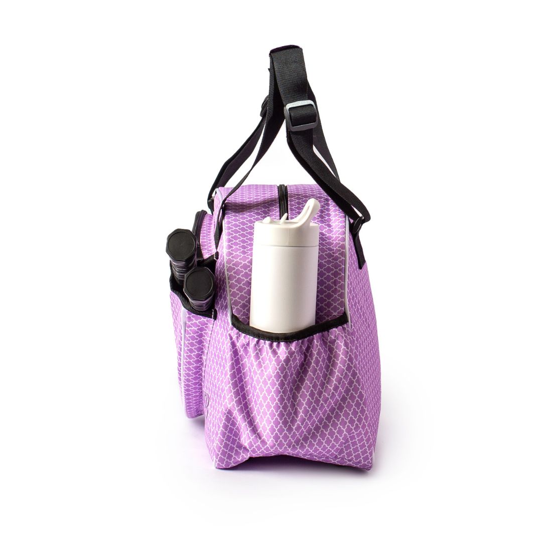 The Pik'le'Ball Sports Classy Premium Women's Pickleball Bag, shown in lavender with a diamond pattern and two outer pockets, features an adjustable black strap. One pocket holds black binoculars, another a white bottle; background is white.