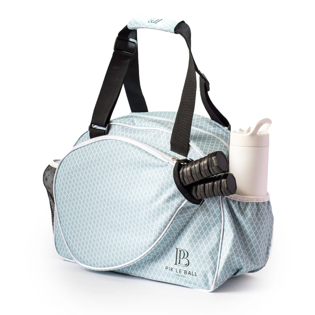 The Pik'le'Ball Sports Classy Premium Women's Pickleball Bag is light blue with a white geometric pattern, black straps, front zip pocket, “Pickleball” logo, and holds three paddles plus a water bottle in its exterior pockets.