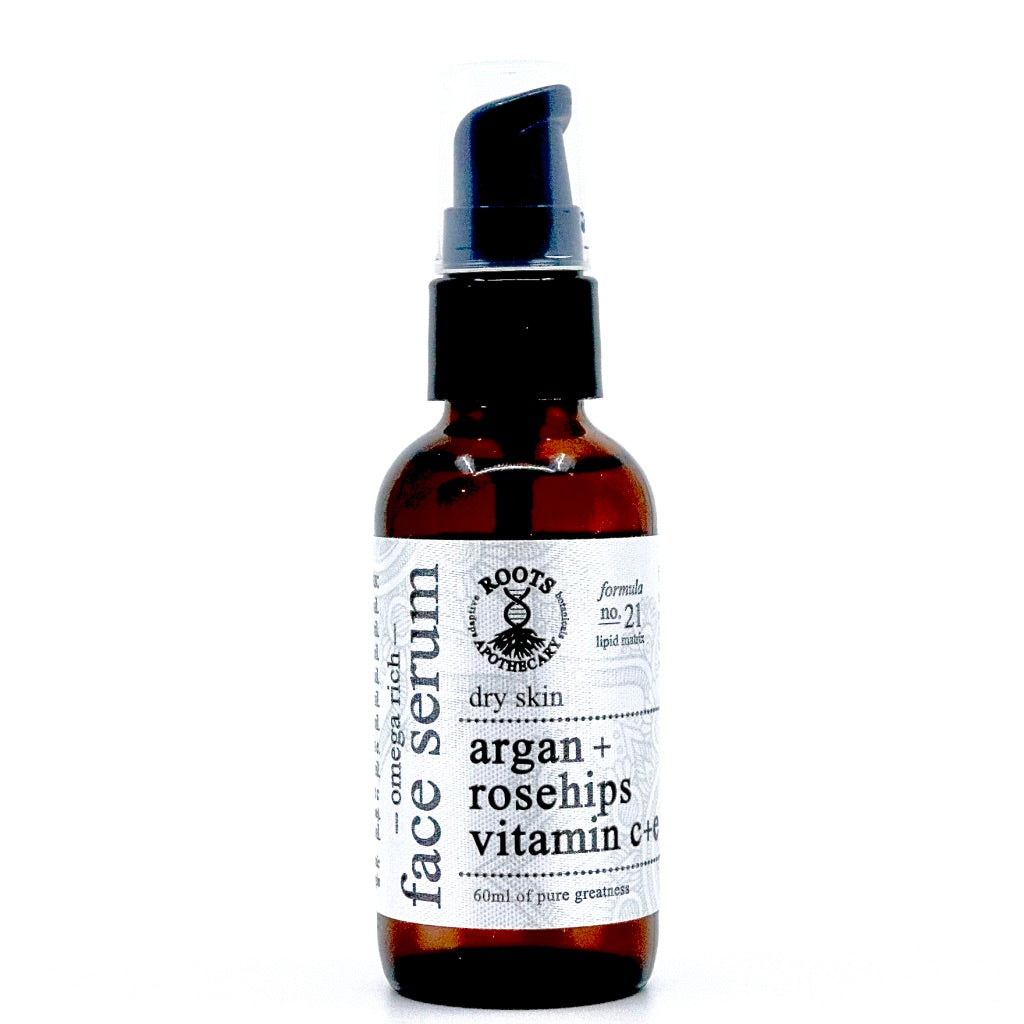 A 60ml brown glass Roots Apothecary face serum for dry skin with a black pump; features unrefined argan oil, rosehips, and vitamins C+E. The white textured label has black text and a circular emblem near the top.