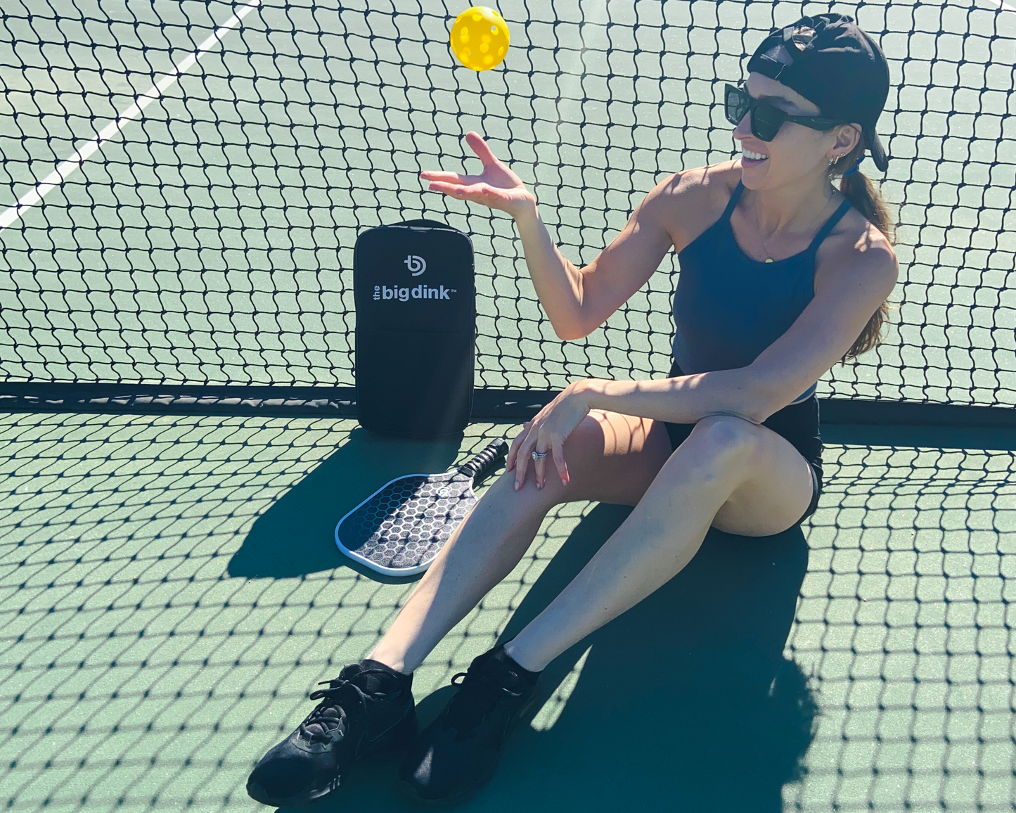 A woman in athletic wear and sunglasses sits on a pickleball court, tossing a yellow ball beside The Big Dink - Model1 Paddle 2-pack Special and a black The Big Dink bag, with the net visible behind her under bright daylight.