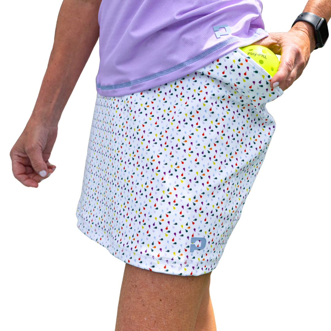 Wearing a lavender top and the Playly Store Sammy Skort 2.0—a court fashion-inspired skort with a colorful geometric print—a person puts a tennis ball in the pocket, sporting a black smartwatch against a plain gray gradient background.