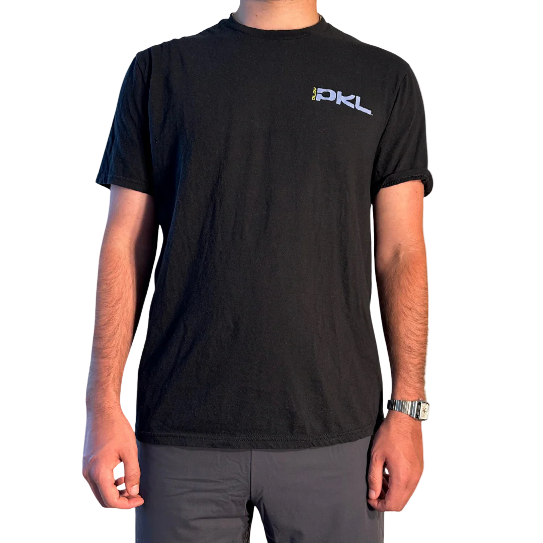 A person wears the Playly Store's Playly - Logo T, a plain black unisex cotton shirt with a small logo on the left chest, paired with gray pants and a silver watch. The photo is cropped at the neck against a plain white background.