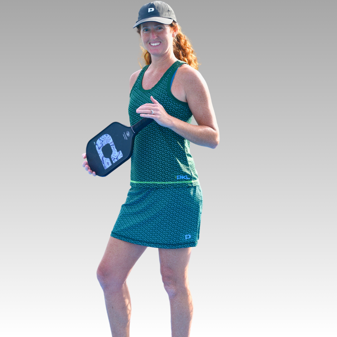 A woman models the Playly Store Sammy Skort 2.0 and a green athletic tank top, smiling with a paddle in hand. She completes her court-ready look with a dark baseball cap and white sneakers against a gray background.