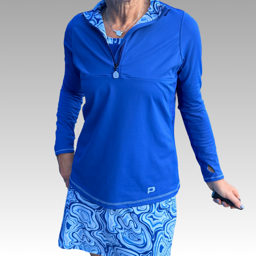 A woman stands against a gray gradient background wearing the Playly Store Soleil Tech Shirt—a vivid blue long-sleeve athletic top with UV protection and zip collar—layered over a patterned tank and matching skirt with wave designs, holding a small dark object.