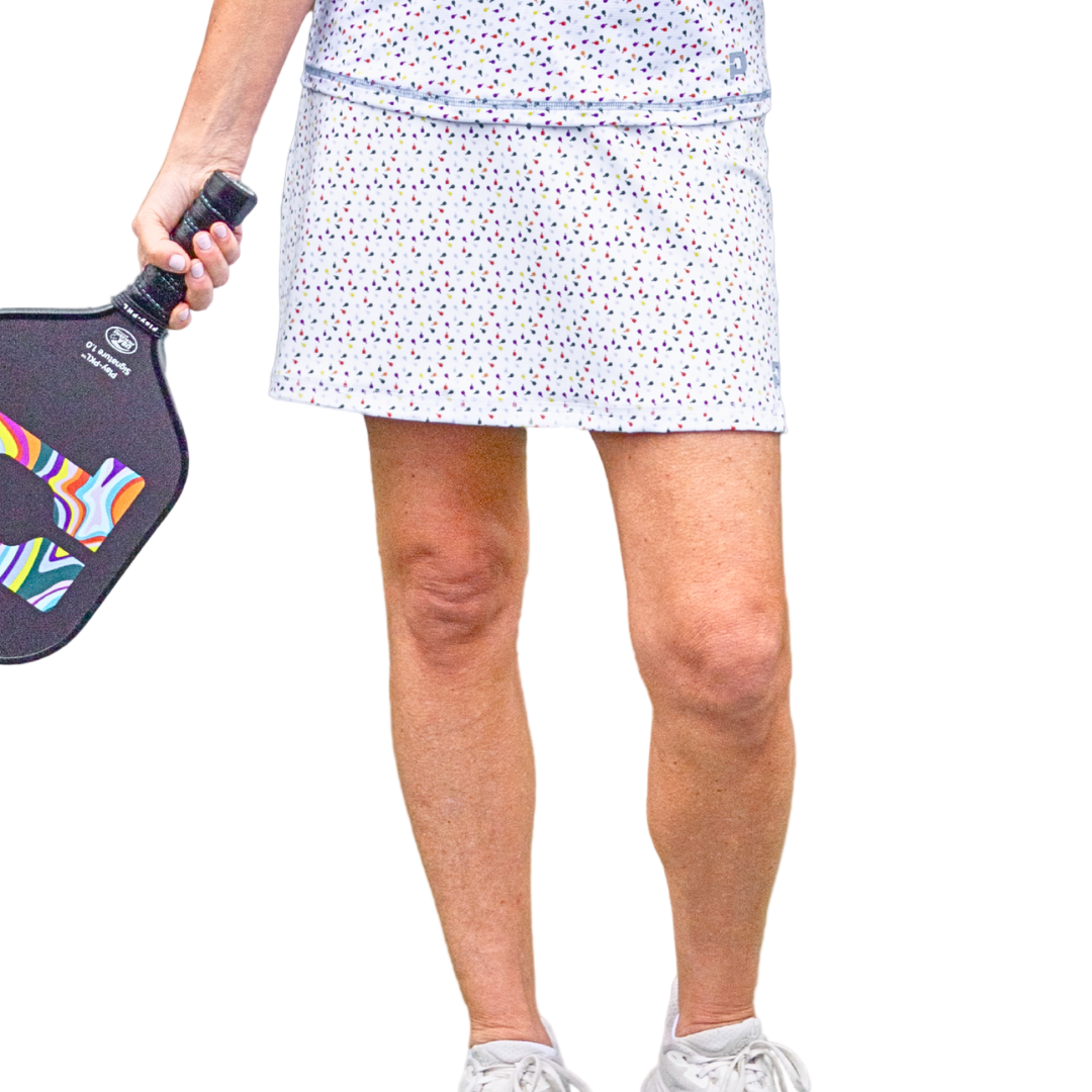 Wearing Playly Store’s Sammy Skort 2.0—a colorful polka dot pickleball skort—and a white top, the figure holds a vibrant paddle, showcasing stylish court fashion and crisp white sneakers against a gray background.