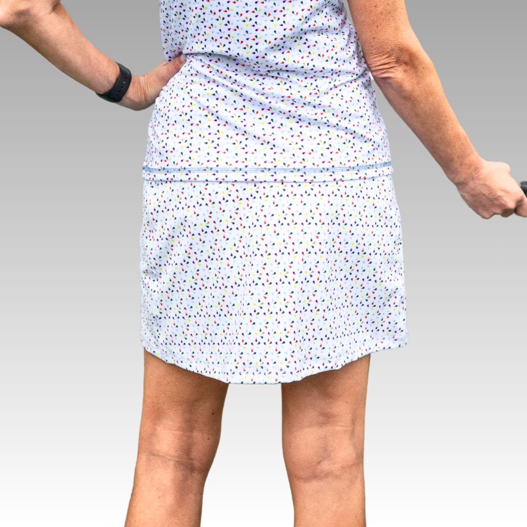 A person stands with one hand on their hip, modeling the Playly Store Sammy Skort 2.0 in light blue with a multicolored confetti pattern and a black watch. The smooth gray background highlights their torso and legs.