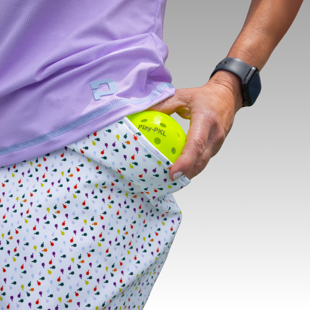 Wearing the Playly Store’s Sammy Skort 2.0 and a purple shirt, a person slips a yellow Play-PKL pickleball into their pocket. A black smartwatch decorates their left wrist, all set against a smooth gray backdrop for effortless court style.