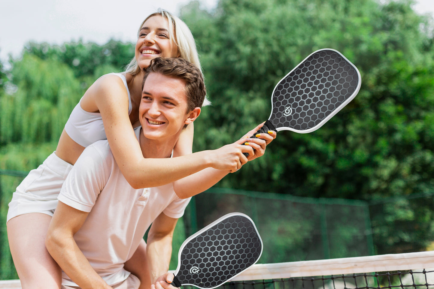 A smiling young woman in white activewear hugs a man in a white polo as they hold The Big Dink Model1 Paddle 2-pack Special by The Big Dink, standing together on an outdoor pickleball court with a net and green trees in the background.