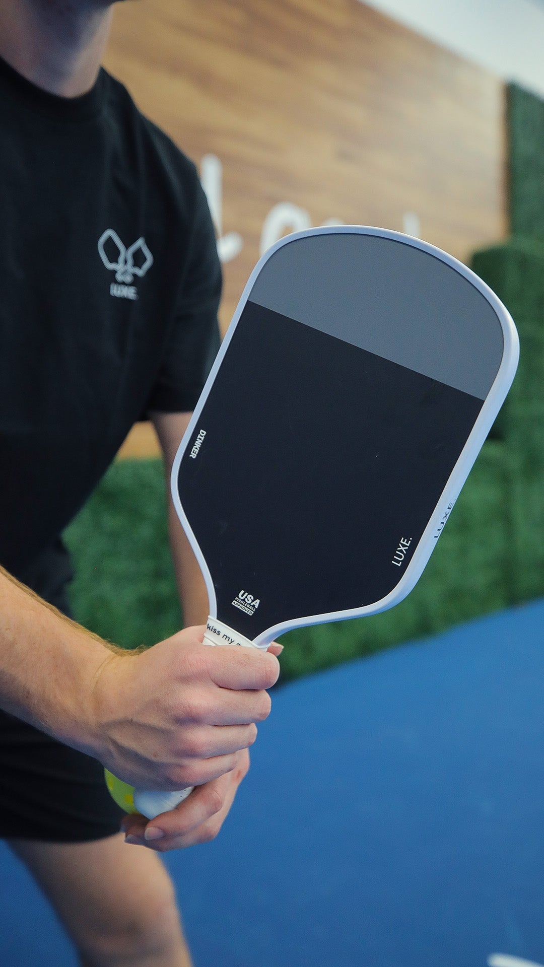A person in a black shirt holds the LUXE Pickleball Dinker carbon fiber paddle, ready to serve. Only their torso, arm, and hand are shown. The paddle’s handle displays USA Pickleball Approved branding; background: green hedge wall and wood panel.