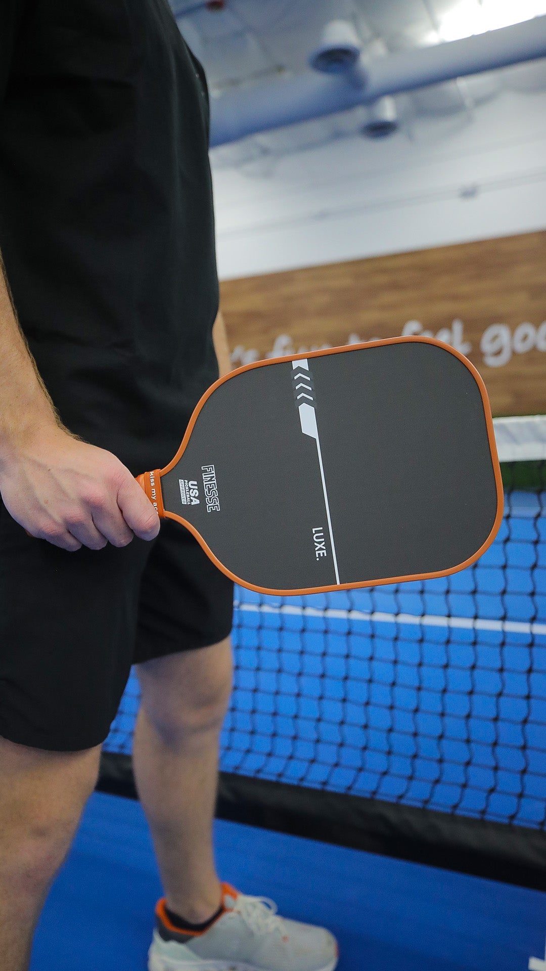 A person in black athletic wear holds a black and orange LUXE Pickleball Finesse carbon fiber paddle on an indoor blue court near the net. Only their lower body and hand are shown; a wooden wall with partial text is behind them.