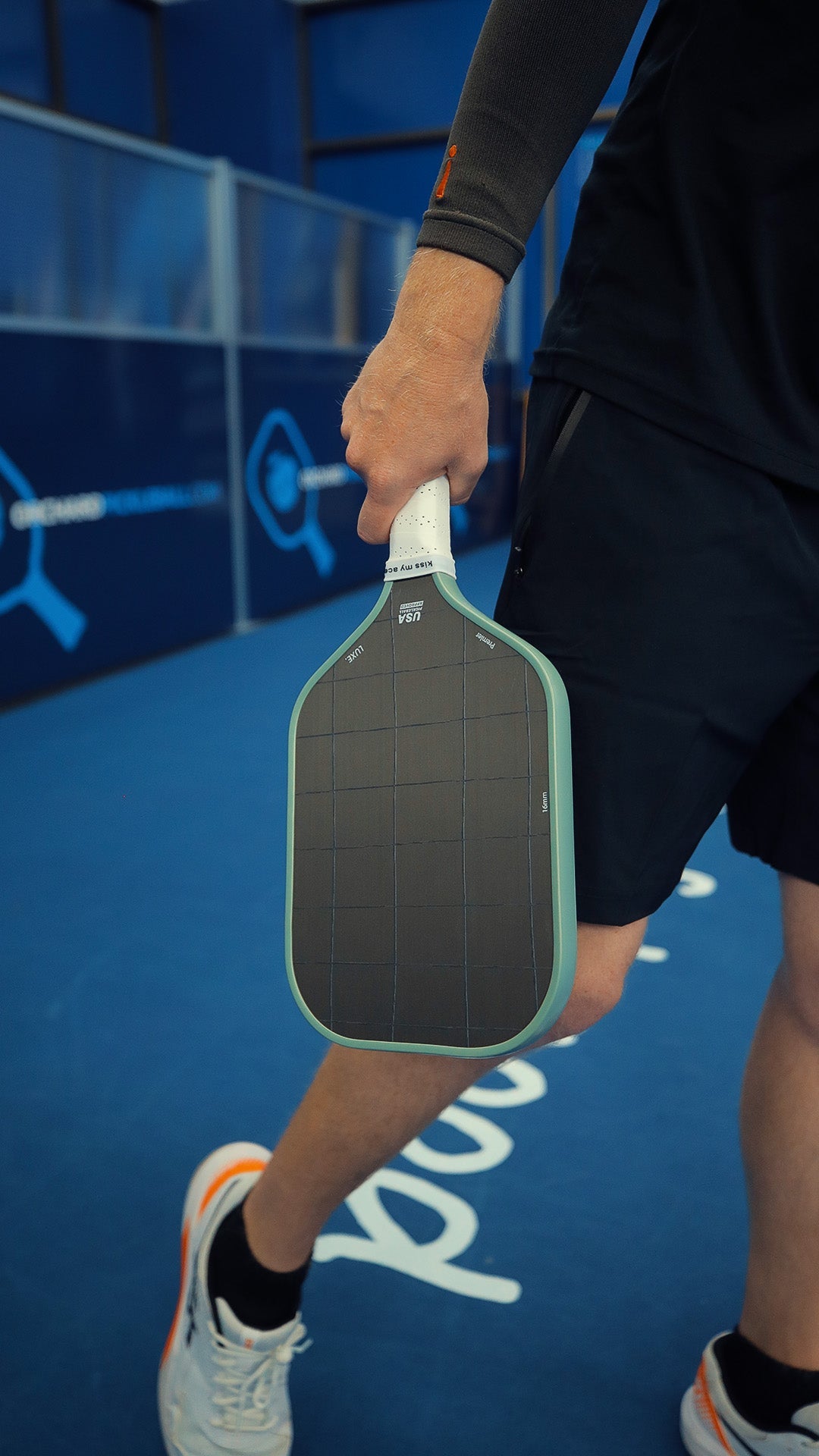 A person in black attire and white sneakers grips the LUXE Pickleball Premier raw carbon fiber paddle on a blue court. Only their lower body and hand are visible, with glass barriers and a court logo in the background.