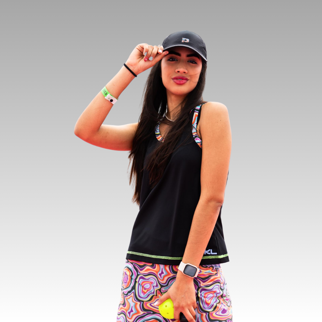 A woman confidently smiles at the camera, wearing the Playly Store Hi Hat Pickleball Hat, a sleeveless black top, and a patterned skirt. She holds a yellow tennis ball and accessorizes with a smartwatch, bracelets, and wristband.