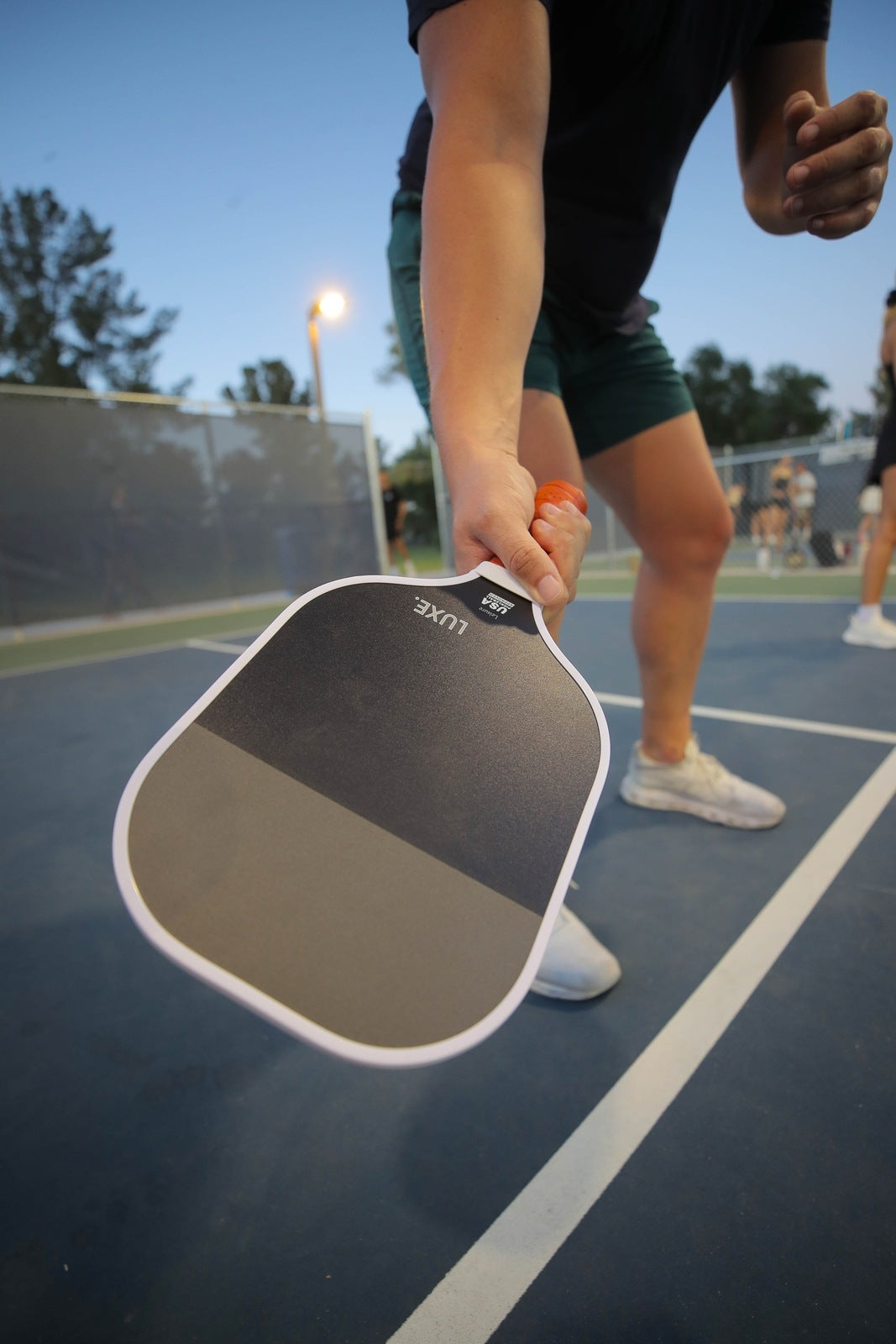 Wearing green shorts and white sneakers, a person holds the LUXE Pickleball Dipped - Leisure paddle with a honeycomb polymer core close to the camera on an outdoor court at dusk, with net and other players blurred in the background.