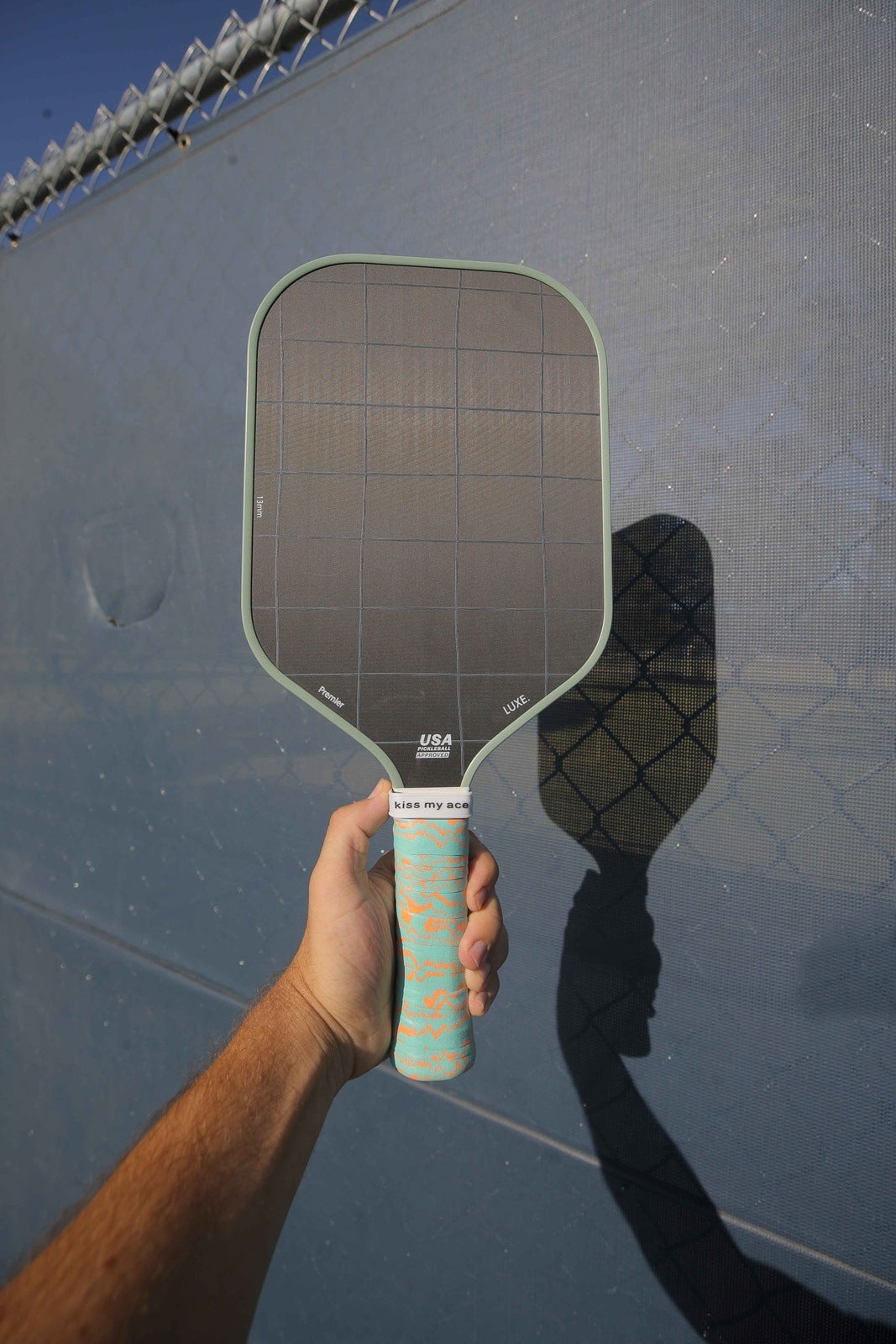 A hand holds a black pickleball paddle featuring a LUXE Pickleball blue Pickleball Paddle Grip and orange designs. Sunlight casts the paddle's shadow on a blue mesh fence with a chain-link fence above.