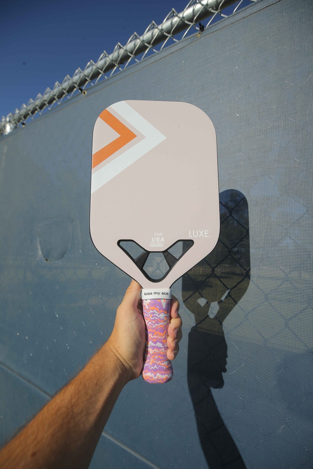 A person holds the LUXE Pickleball Draft paddle with a pale pink face, orange and white chevrons, "LUXE." branding, and a wavy-patterned grip. Blue and chain-link fences appear in the sunny background.