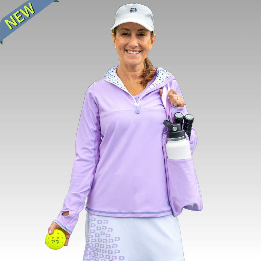 A smiling woman wears the Playly Store Soleil Tech Shirt and a white cap, holding a yellow pickleball. She carries paddles and a water bottle in her pocket. A “NEW” banner is displayed on the gray background.