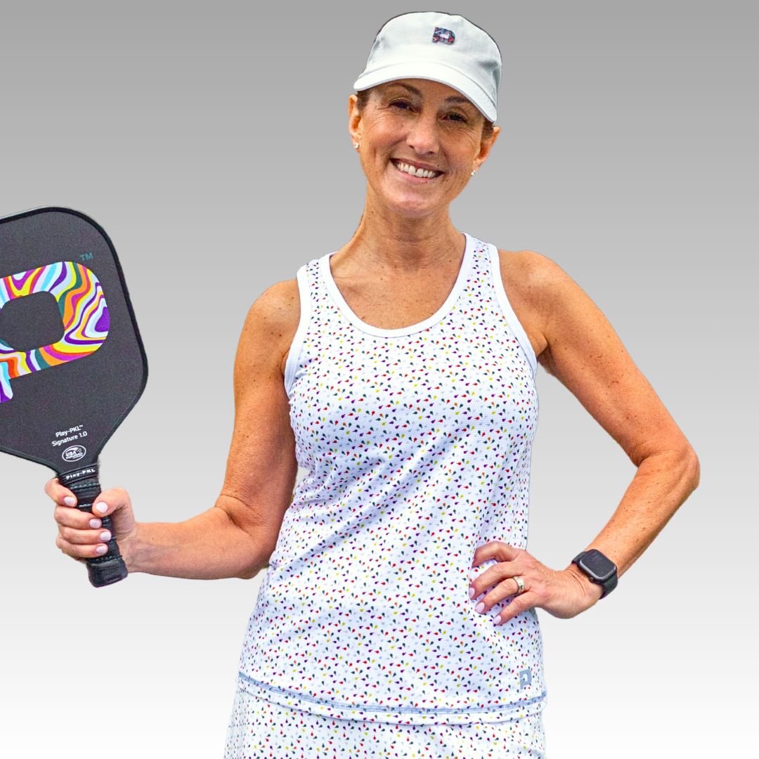 A smiling woman in the Playly Store Pretty Tank—a colorful dot-patterned, moisture-wicking athletic mesh top—pairs it with a matching skirt and vibrant paddle, confidently posing against a gray gradient background.