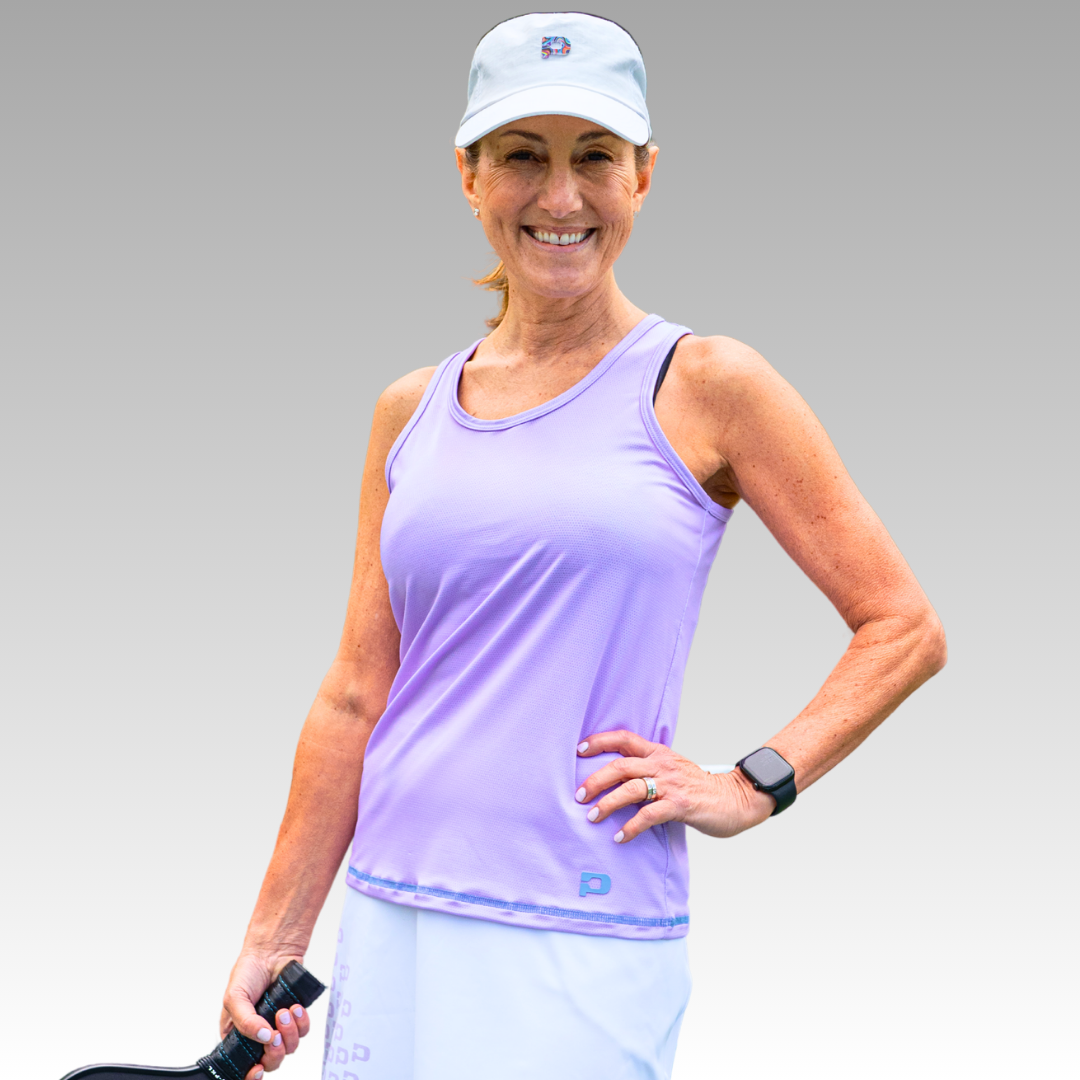 A woman smiles confidently in the Playly Store Pretty Tank, a lavender moisture-wicking pickleball top, paired with a white cap, white skirt, and smartwatch. She stands with one hand on her hip, holding a pickleball paddle against a gray background.
