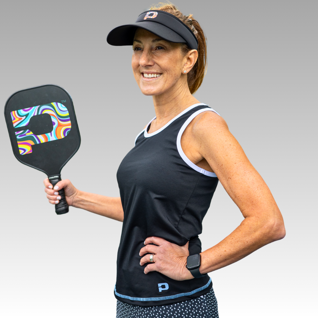 A woman smiles, holding a colorful paddle. She sports a black visor, Playly Store’s Pretty Tank with white trim, a patterned skirt, and smartwatch. Her hair is in a ponytail against a smooth light gray background.