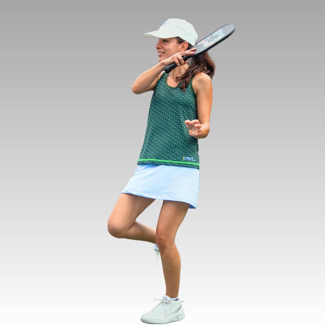 A woman in the Playly Store Pretty Tank (green) pairs it with a light blue skirt, white sneakers, and a white cap as she plays tennis mid-swing with a black racket against a plain gray gradient background.