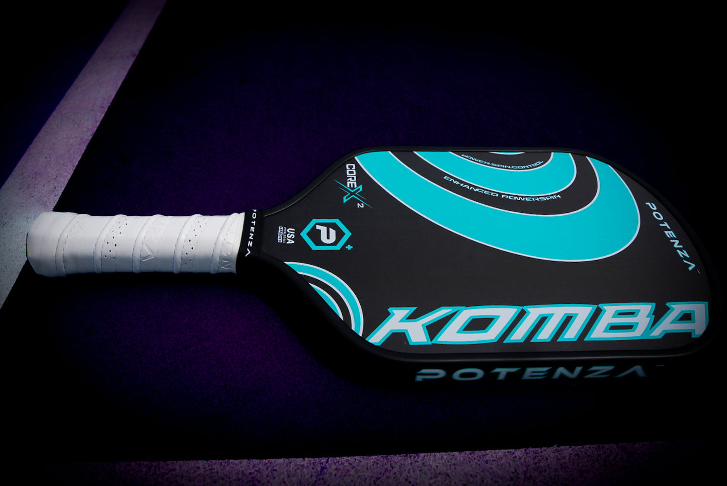 A black carbon fiber KOMBA+ COREx2 paddle from Potenza Pickleball, with teal and white accents, a white grip, and geometric designs lies on a dark purple court near a white boundary line.