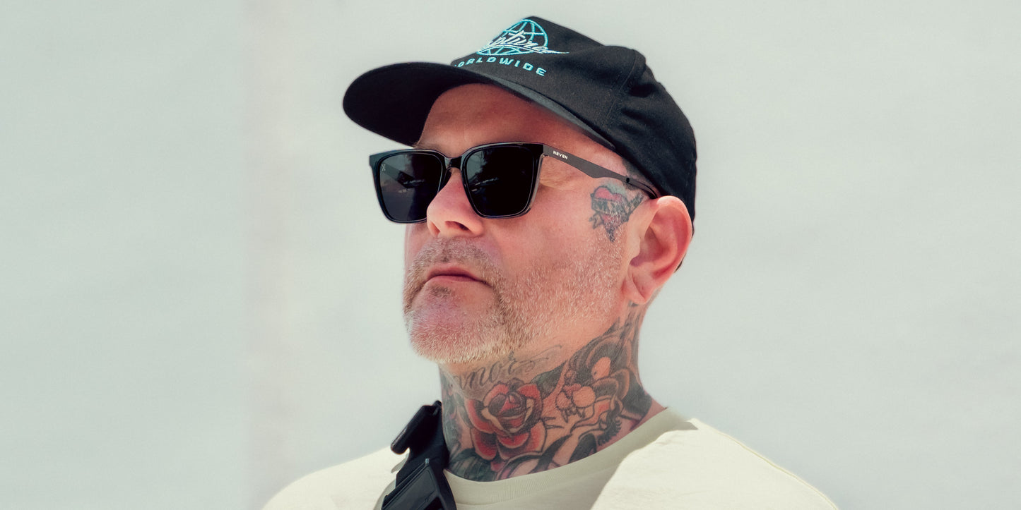 A man with tattoos on his neck and face wears Neven Eyewear Mamba polarized sunglasses with black frames, a black cap, and a light shirt. He stands against a plain background with a calm expression and carries a black strap.