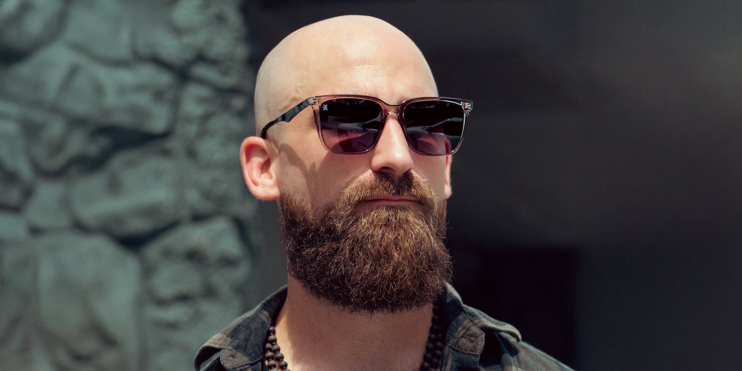 A bald man with a thick brown beard wears Neven Eyewear's Tornado sunglasses and a dark shirt. Standing outside in bright sunlight before a blurred stone wall, he looks neutral and confident.