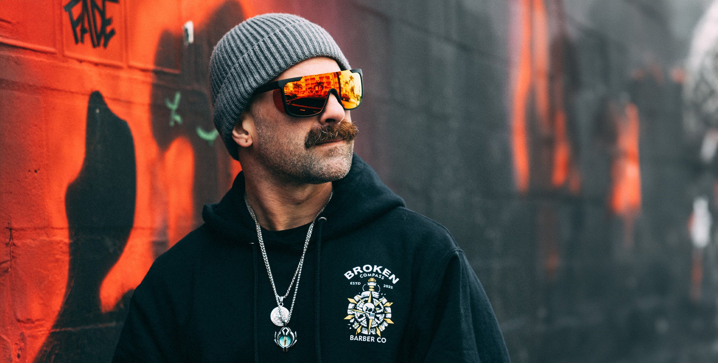 A man with a mustache in a gray beanie and black Broken Barber Co. hoodie wears Neven Eyewear Blood Moon sunglasses with impact-resistant lenses, standing against a graffiti wall featuring bold red and black street art.