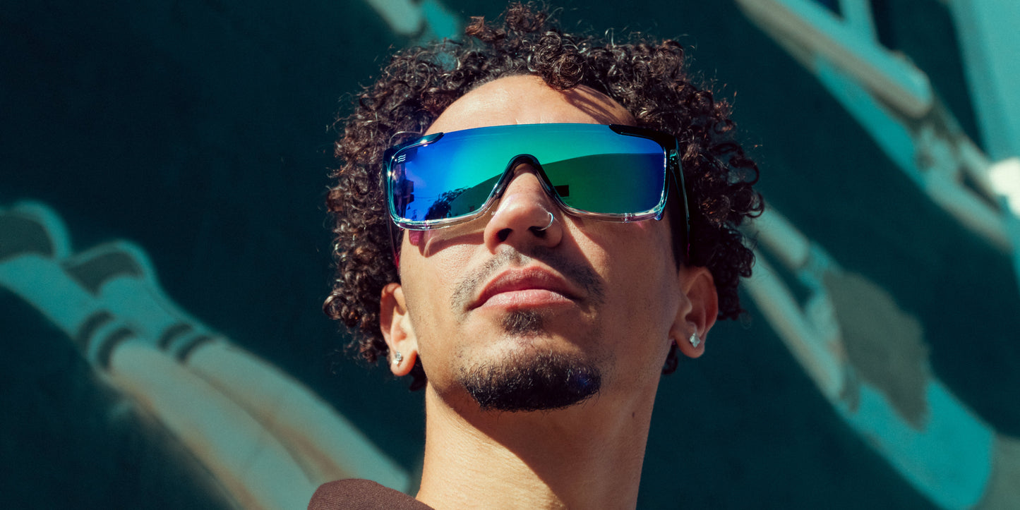 A person with short, curly hair and facial hair looks up, wearing oversized mirrored Neven Eyewear Night Watch sunglasses with removable side shields from the Daywalkers Collection. Behind them is a blue-green mural with abstract shapes and pale figures.