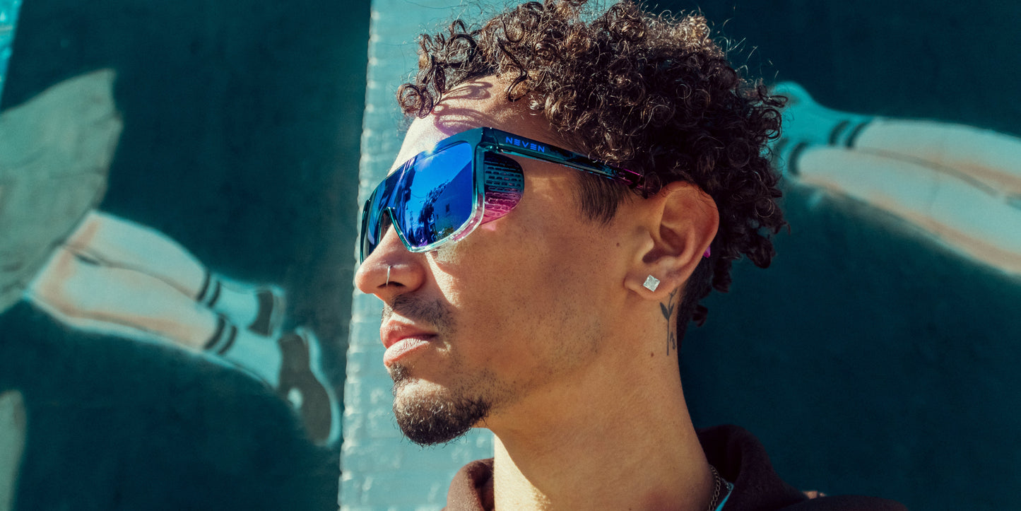 A man with short curly hair, a goatee, and wearing Neven Eyewear's Night Watch sunglasses gazes to the side. He stands before a mural of legs in striped socks on a dark green background as sunlight illuminates his face.