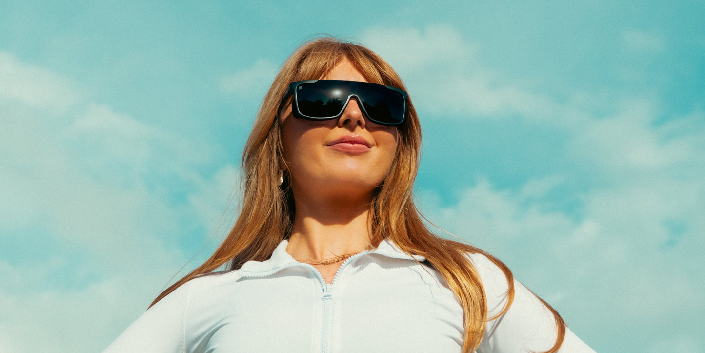 A confident woman with long, straight blonde hair wears Neven Eyewear’s Pitch Black sunglasses with removable side shields and a white zip-up top, smiling outdoors beneath a bright blue sky with scattered clouds.